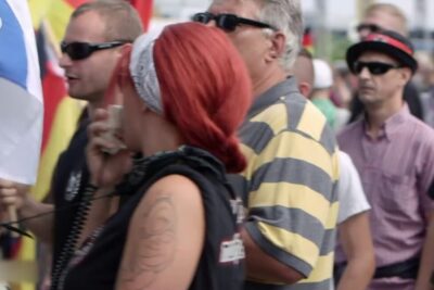 A red-haired organizer speaks into a megaphone amid a crowded street rally, evoking themes of commercialisation excessive.