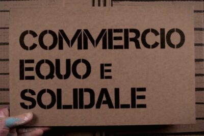 Brown cardboard sign stenciled with COMMERCIO EQUO E SOLIDALE held by a hand, commercio equo 1