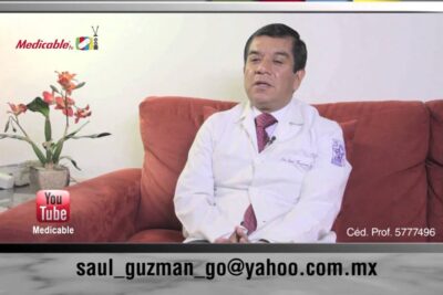 Medical professional in white coat seated on a red couch during a filmed interview about complicaciones embarazo 1.
