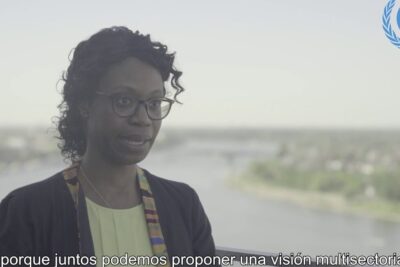 Woman wearing glasses and patterned scarf speaking on a riverside balcony with Spanish subtitles, comunidades locales 1