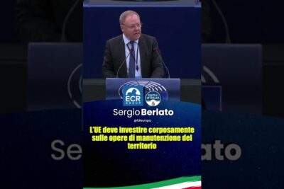 A suited speaker at a podium in a formal assembly calling for investment in comunita locali infrastructure maintenance