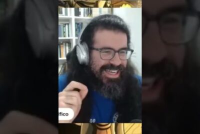 Smiling person with long hair, beard, glasses and headphones in front of a bookshelf, corriente deterministe