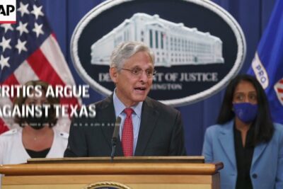 Senior official speaking at Department of Justice podium with U.S. flags and masked aides as critics argue 1.