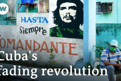 Man in a face mask seated beside a colorful wall mural with a stenciled revolutionary portrait and painted slogan referencing the cuban regime.