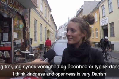 A woman being interviewed on a narrow city street with subtitles referencing danske sprog
