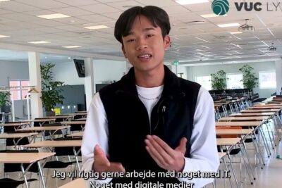 Young student speaking in a large study hall with rows of desks and Danish subtitles about IT, danske uddannelsessystem 1