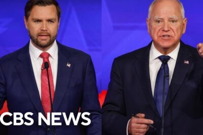 Two men in suits standing at podiums speaking during a televised debat tsar event
