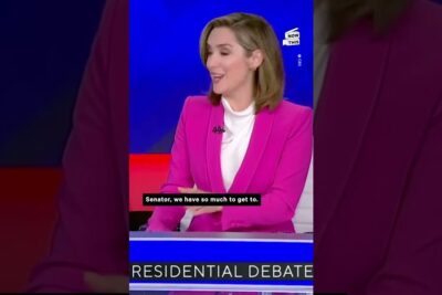 Television moderator in pink blazer seated at a news desk during debate racismo 1.