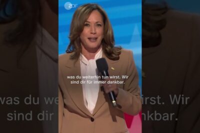 Woman in a tan blazer speaking into a handheld microphone on stage with German subtitles visible, debatten lederskap.
