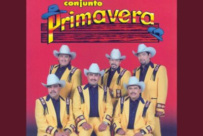Seven-member conjunto wearing matching yellow suits and white cowboy hats against a red background, derecho vida 12.