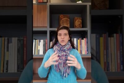 Woman in a blue sweater and multicolored scarf gesturing in front of a bookshelf while discussing desacordo trabalhista 2.