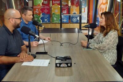 Three people seated around a podcast table with microphones and a small radio, each desempenha papel in the discussion