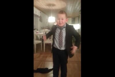 Child wearing a suit and tie standing mid-gesture in a warmly lit dining room, det ar 33.