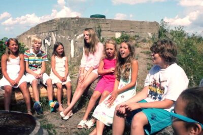 A group of children sitting outdoors on logs and rocks singing together at a sunny location det ar 7.