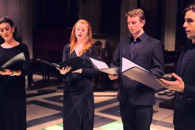 Four singers in black formal attire hold open sheet music and perform as a focused vocal quartet in a lit hall, det ar 79.