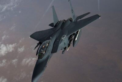 A twin-engine tactical fighter jet flying above scattered clouds and desert terrain with visible cockpit and external fuel tanks, det er 27.