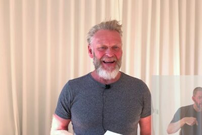 Smiling bearded presenter with lapel mic and paper in front of beige curtains, det vigtigt 18