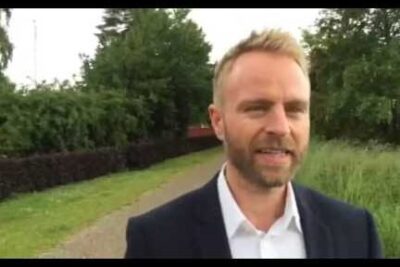 Adult man in a dark blazer and white shirt walking along a grassy tree-lined path, det vigtigt 6