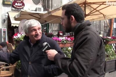 An older man being interviewed on a city street near a café about diritti donne