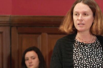 Woman speaking at a formal event with wooden paneling background about discrimination positive