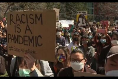Masked protest crowd with a cardboard sign reading 'RACISM IS A PANDEMIC' highlighting discrimination raciale 1
