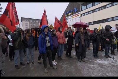 A wintertime crowd in front of a commercial building holding red flags and umbrellas during a street demonstration, economique mondial 1.