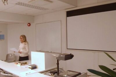 Woman presenting in a classroom with whiteboard and projector holding papers during eettisia kysymyksia 6