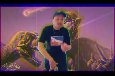 Person in a cap and graphic tee poses in front of two large dinosaur figures with neon glitch effects and bold colors, ein weiterer 11.
