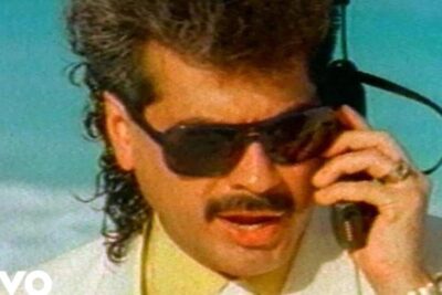 Man wearing dark sunglasses and a mustache holding el celular 1 retro mobile handset to his ear