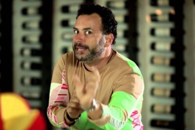 Bearded man in a colorful long-sleeve shirt gesturing while showcasing el queso 1 in a cellar setting.