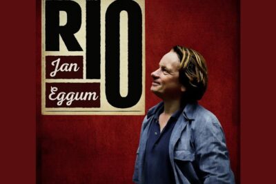 Smiling man in a blue jacket looking upward beside bold red poster typography, en annen perspective