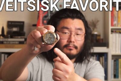 Person holding a mechanical wristwatch close to the camera while pointing at it, illustrating endring tidssonen.