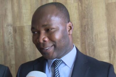 Person in a dark suit and striped tie speaking into a microphone during an interview about entreprises publiques