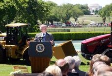 Suited speaker at an official podium on a sunny lawn with construction machinery and a domed memorial, highlighting equidad