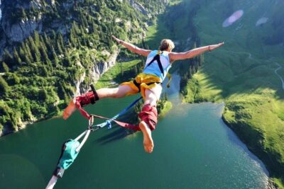 A person bungee jumping from a cliff over a green alpine lake wearing a harness and ankle straps esportes extremos 1.