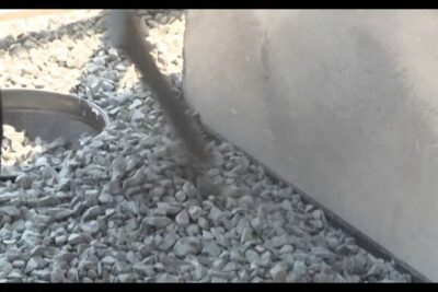 Close-up of gray gravel beside a concrete curb and a metal access ring with a shadow, esteettisiin tarkoituksiin 1.