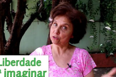 Woman in a pink floral top speaking outdoors beside trees and plants, gesturing as she explains how to estimular imaginacao