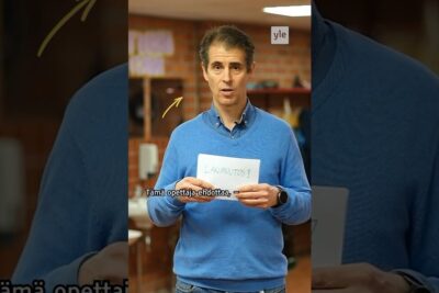 A teacher in a blue sweater holds a handwritten card in a classroom with Finnish subtitles and a Yle logo etta koulukiusaaminen 1