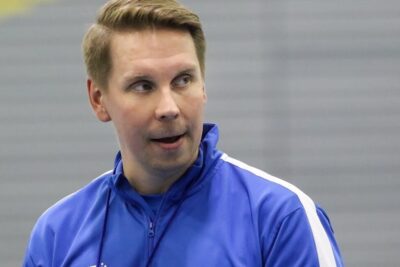 Coach in a blue tracksuit attentively observing players during indoor etta pelaaminen practice.