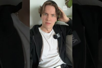 Young person in a black hoodie and white tee sitting on a sofa with a lapel microphone and hand in hair, etta tiettyjen.