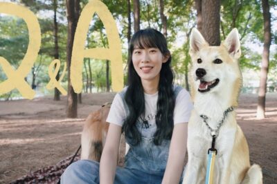 A smiling person sits on a blanket in a tree-lined park beside a leashed dog, eutanasian kriteerit 1