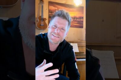 Smiling person in a cozy wooden home studio with a ukulele on the wall and piano sheet music, labeled existenz gottes 2