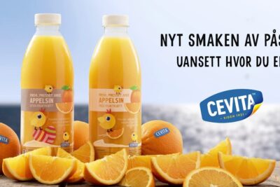 Two bottles of ferskpresset appelsinjuice with whole and sliced oranges on a wooden surface by the water