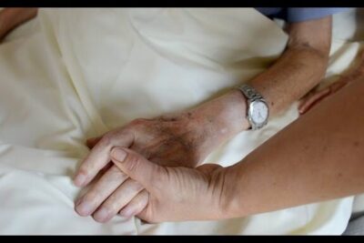 Two hands gently holding an older adult's hand on a white bed sheet, fin vie 1.