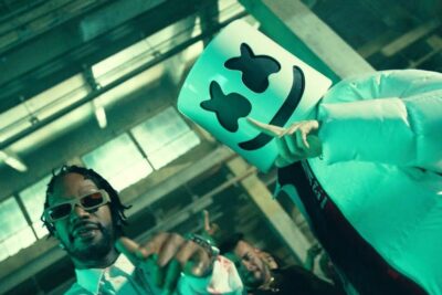 Two performers in a green-lit industrial space, one wearing a smiling marshmallow-style mask and the other in sunglasses for hitta.