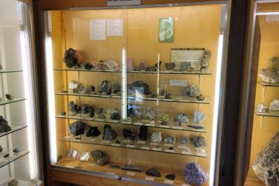 Glass-fronted cabinet of labeled Norwegian rock and mineral specimens on glass shelves with vertical lighting, fore til 16