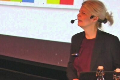Female presenter with headset microphone on stage delivering a talk forstaelse varlden