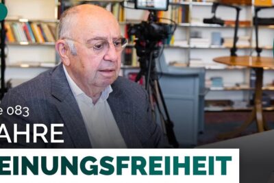 Elderly man seated in a media studio with cameras, bookshelves and a laptop illustrating frei aussern and public discussion.