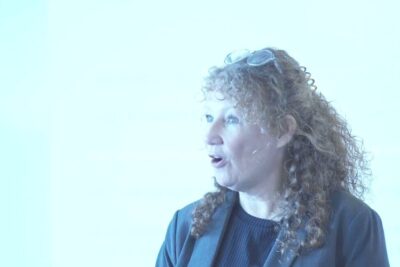 Curly-haired instructor with glasses on her head speaking animatedly in a bright room for fysisk undervisning 1