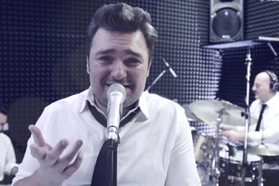 Male lead vocalist at a microphone with a drummer and keyboardist in a padded studio performing a lively genere musicale.